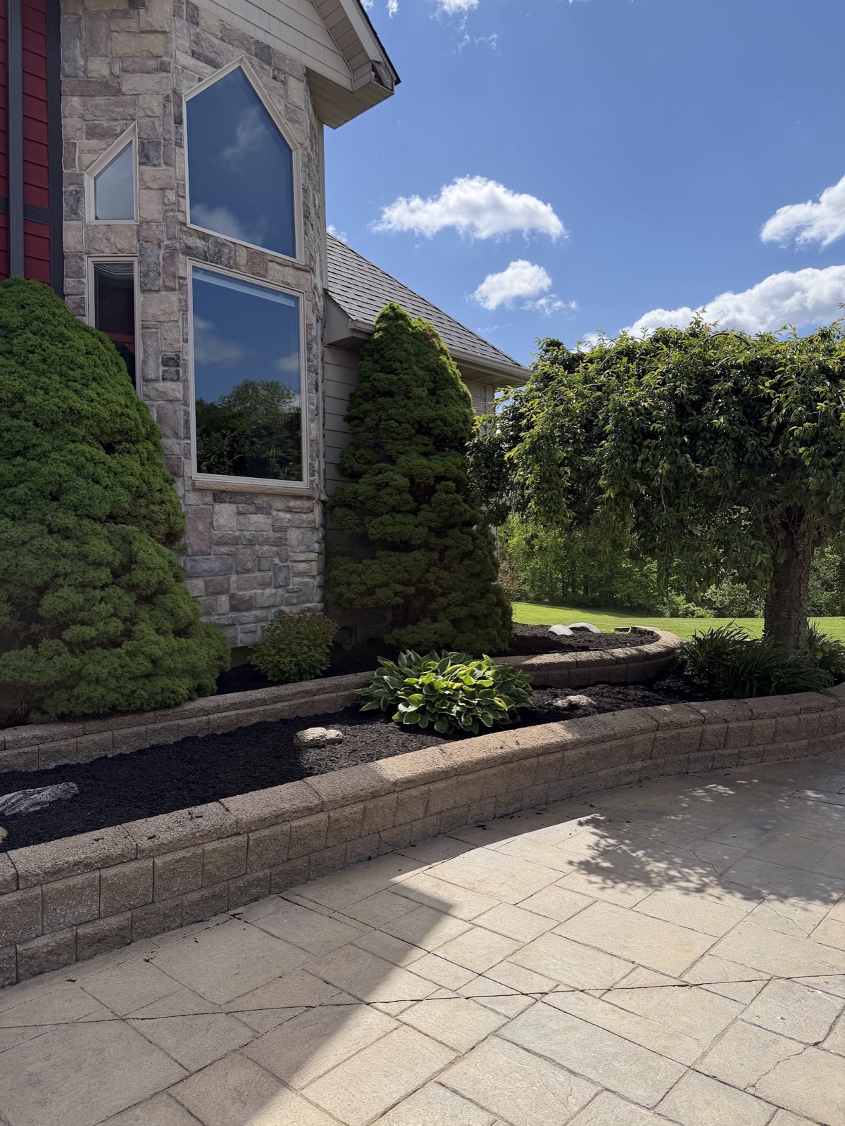 Curved block retaining-wall beds with fresh black mulch and hostas beside a stamped concrete patio at a stone-front home.