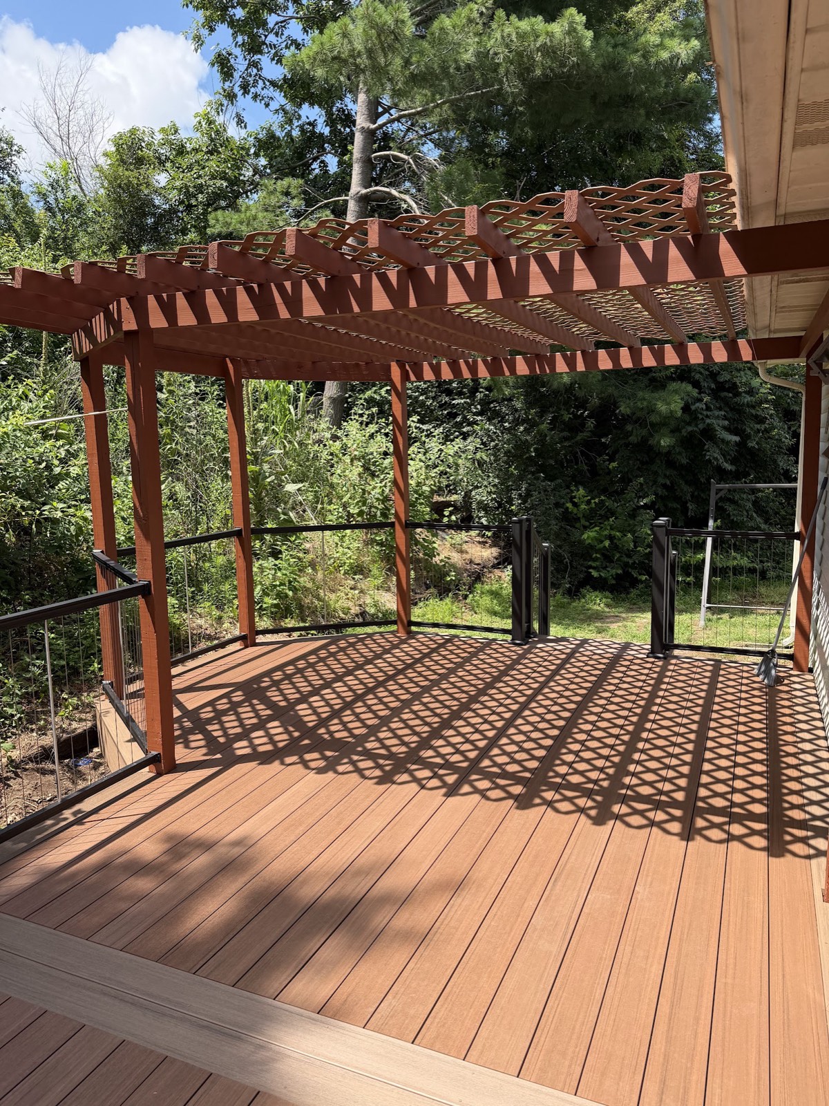 Red-stained pergola with lattice top over a spacious new composite deck with black cable railing.