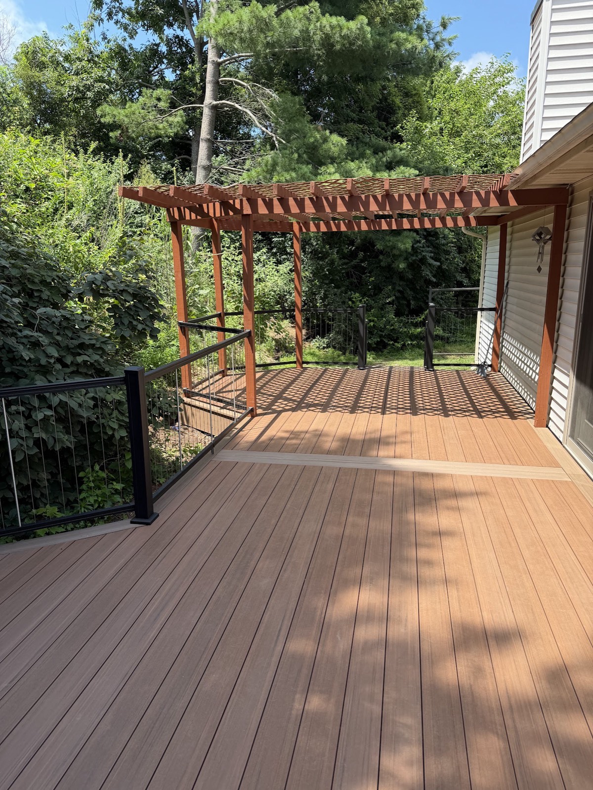 New composite deck with breaker board detail, black cable railing, and a red-stained pergola casting lattice shadows.