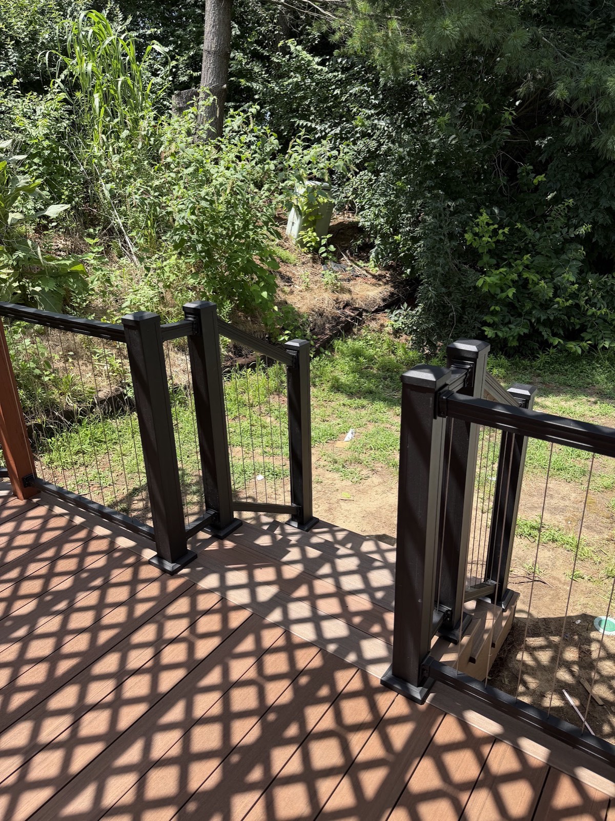 Detail of black vertical-cable deck railing and stair opening on new composite decking with lattice shadows.