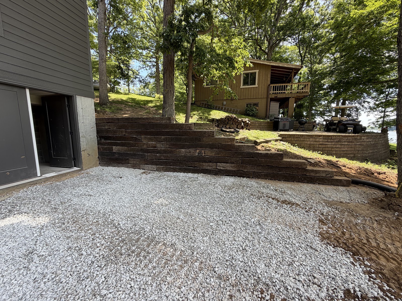 Tiered railroad-tie retaining wall and fresh gravel pad built into a wooded hillside below a lake cottage.