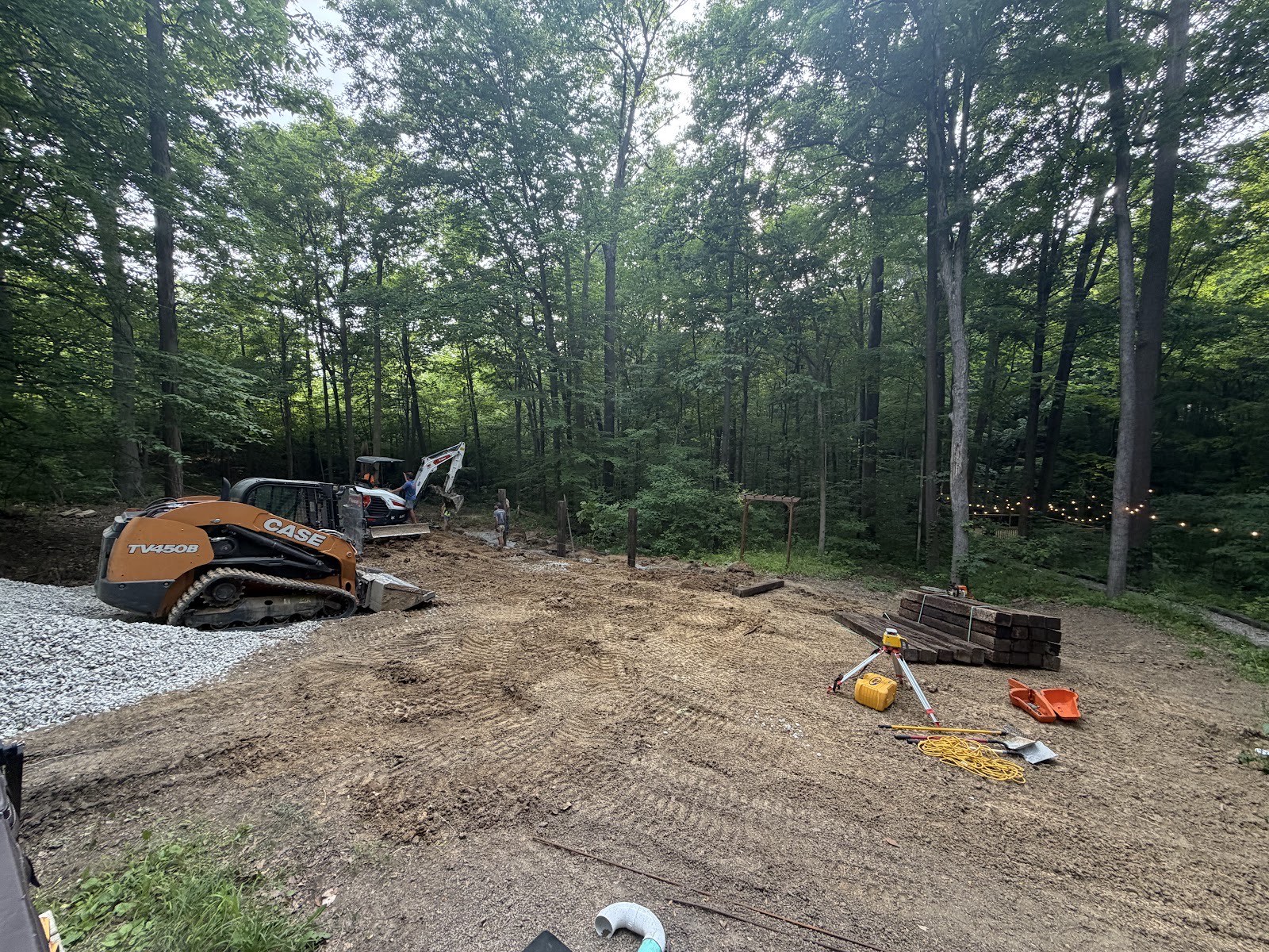 CASE track loader and mini excavator grading dirt in a wooded backyard with stacked railroad ties and a laser level.