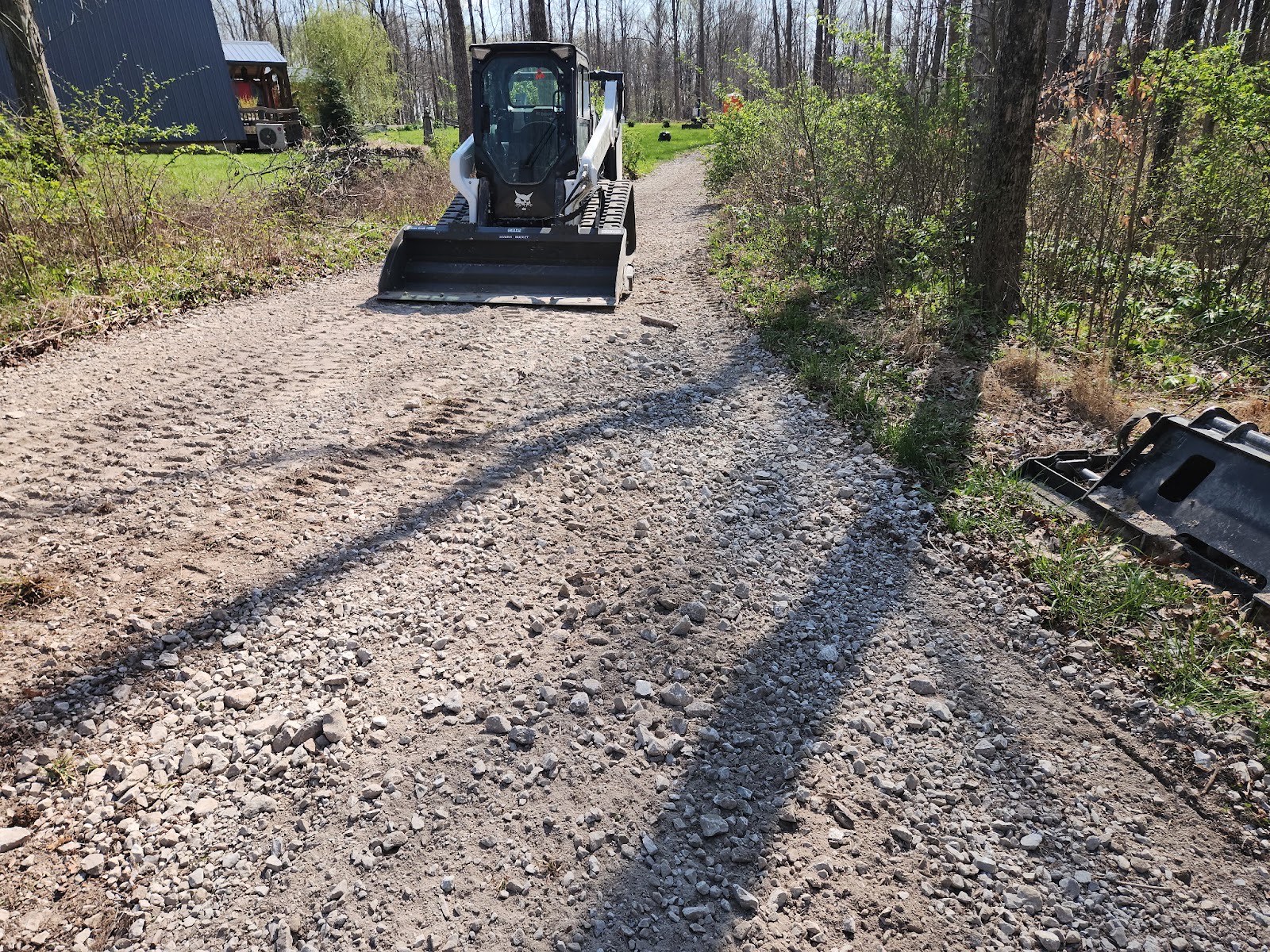Bobcat track loader spreading and grading fresh gravel on a wooded driveway.