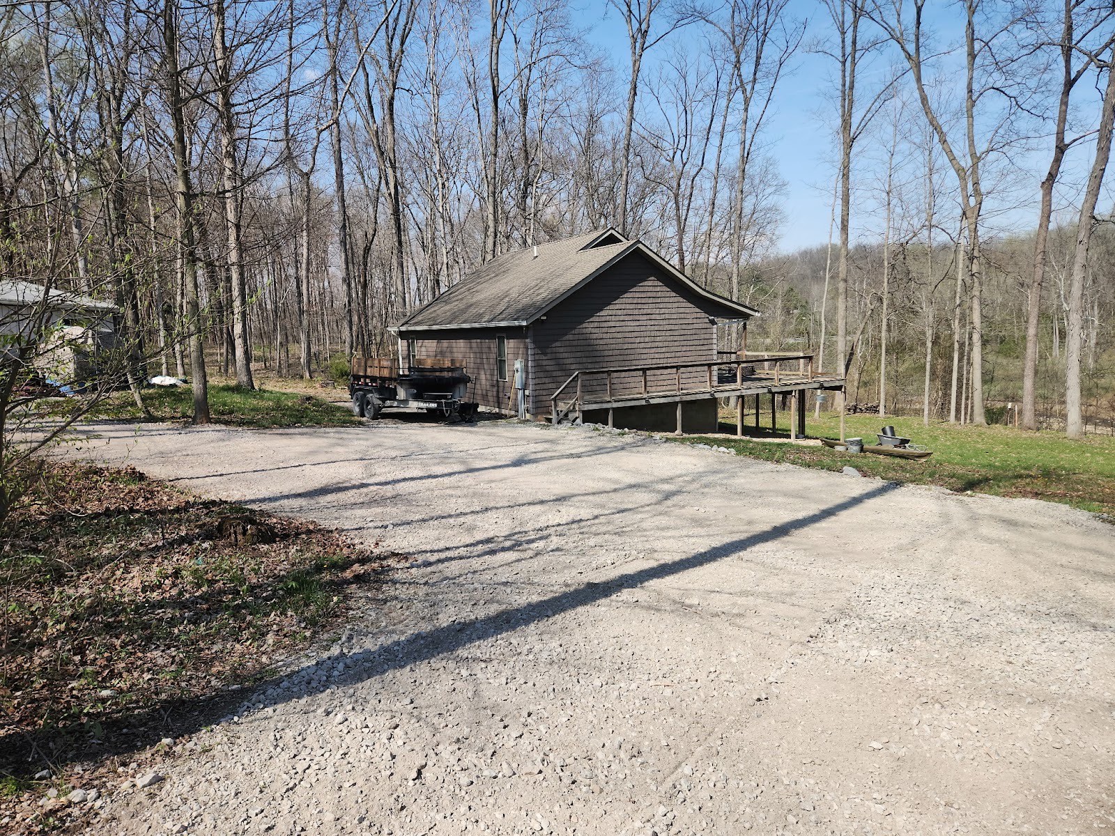 Freshly spread gravel driveway and parking area at a wooded cabin with an empty equipment trailer nearby.