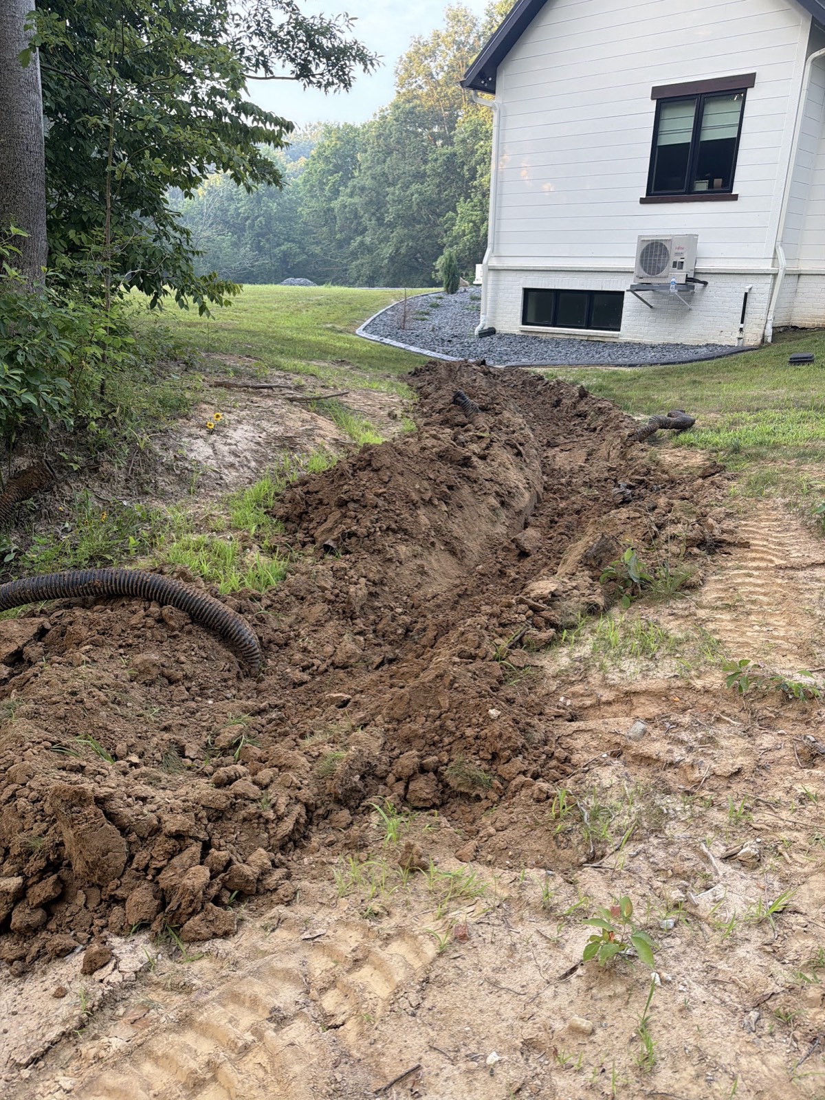 Open drainage trench with corrugated pipe and mounded dirt running toward a white brick house.