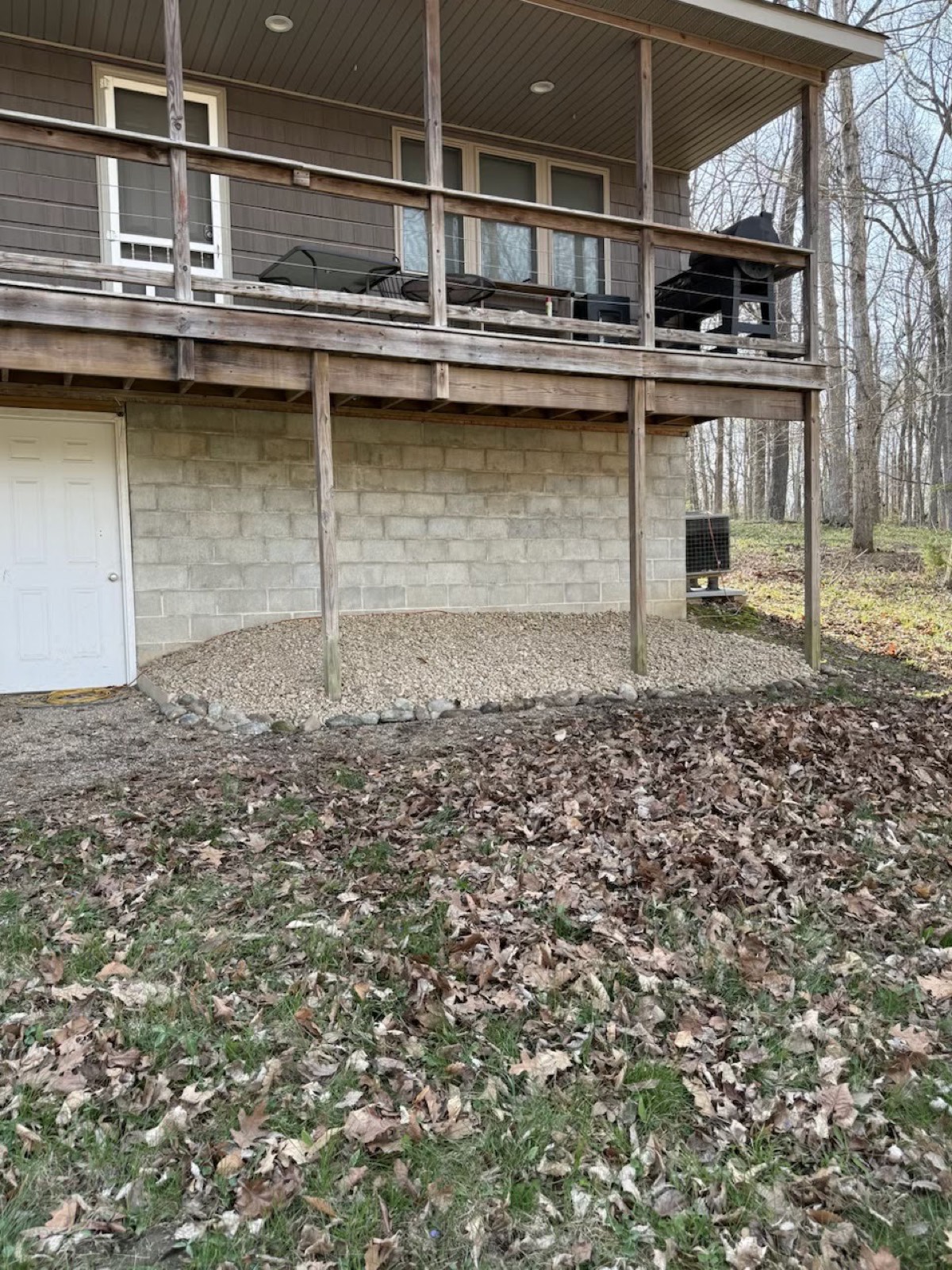 Leaf-covered yard below an elevated wooden deck with a gravel bed against the block foundation.