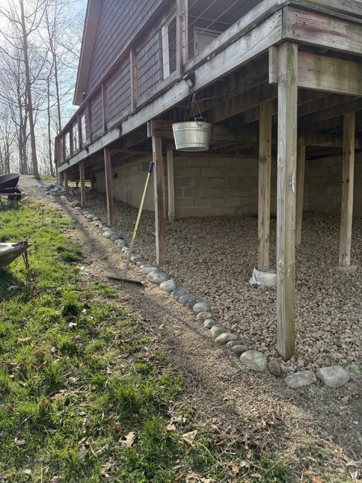 New gravel drainage bed with river-rock border installed beneath an elevated wooden deck on a slope.